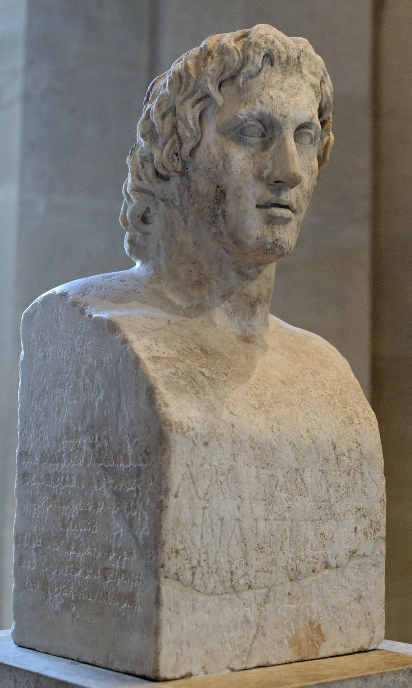 Weathered marble herm bust of a young man with thick tousled hair, the nose damaged, inscribed on its base